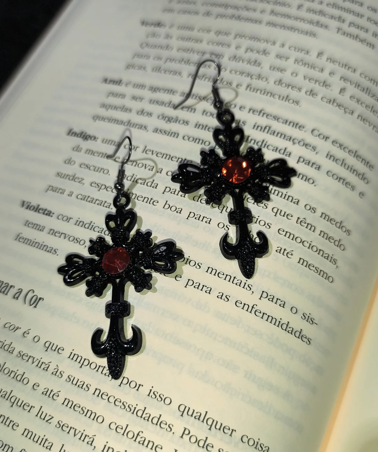 Brinco cruz preta vermelha gótico em liga metálica com detalhes em flor-de-lis e strass vermelho central sobre páginas de um livro.