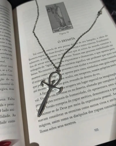 Colar ankh espada gótico com corrente de aço e pingente de liga metálica posicionado sobre a página do livro O Eremita.