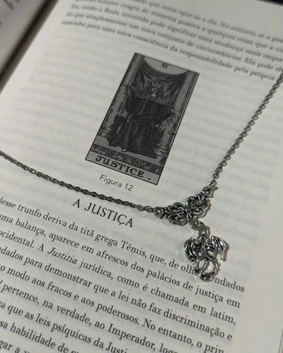 Colar dragão gótico com corrente de aço e pingente detalhado sobre a página de um livro aberto com ilustração de tarô.