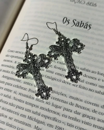 Brinco cruz com mariposa gótico e caveira em liga metálica prateada envelhecida sobre páginas de um livro.