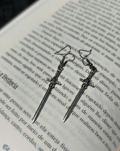 Brinco espada gótica medieval em liga metálica prateada envelhecida com empunhadura detalhada sobre páginas de um livro.