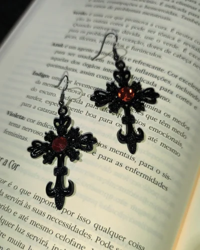 Brinco cruz preta vermelha gótico em liga metálica com detalhes em flor-de-lis e strass vermelho central sobre páginas de um livro.