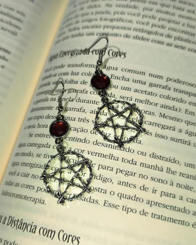 Brinco pentagrama místico vermelho gótico em liga metálica prateada texturizada sobre as páginas de um livro.