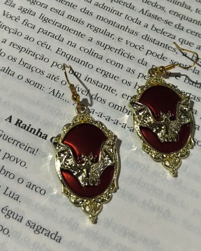 Brinco morcego camafeu vermelho gótico em liga metálica dourada sobre as páginas de um livro